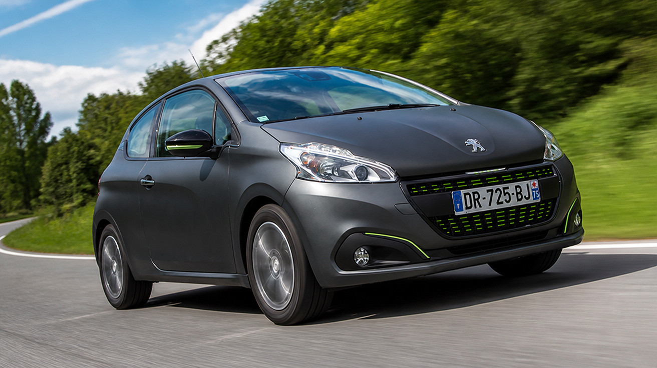 Matte black Peugeot compact hatchback driving on a curved road with green trees and blue sky background.