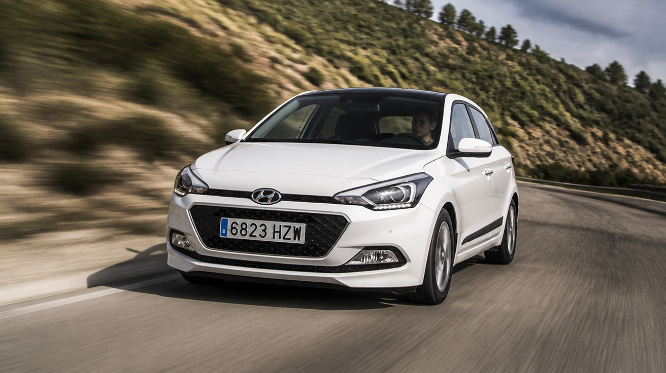 White Hyundai car driving on a winding road with grassy hills and trees in the background.