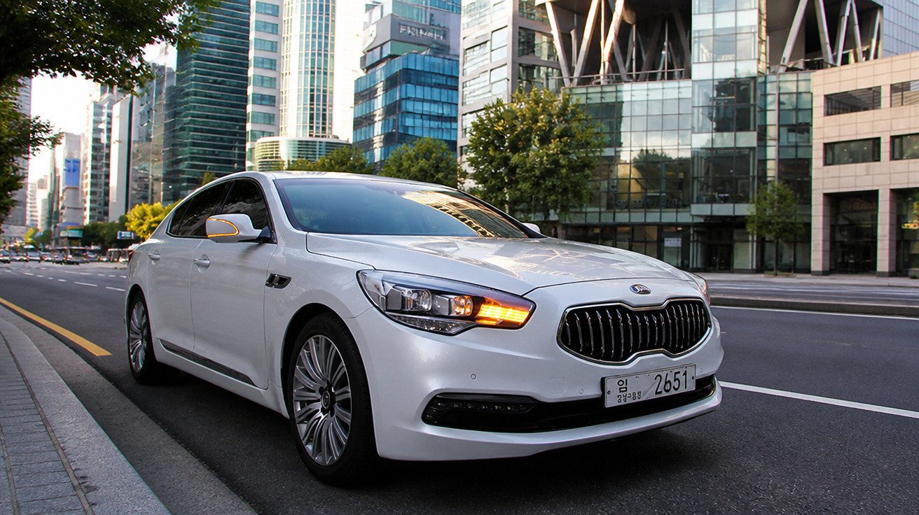 White Kia sedan with headlights on parked on a city street beside modern glass buildings.