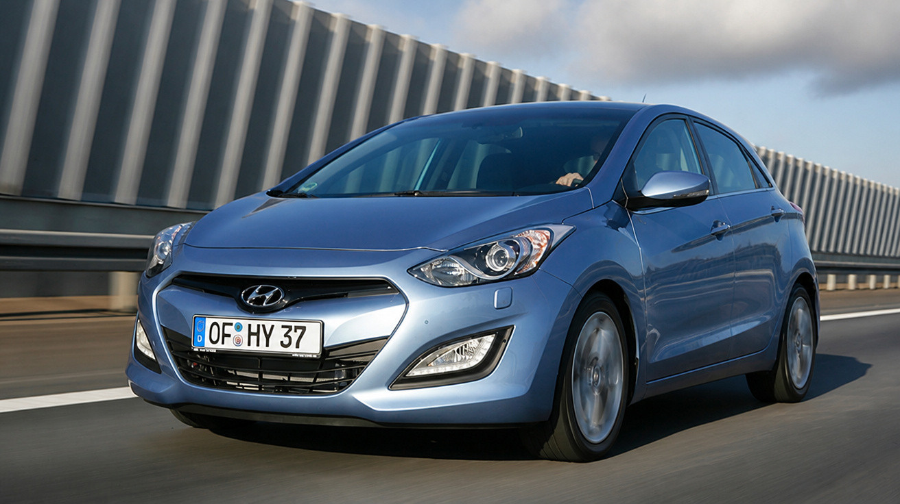 Blue Hyundai hatchback driving on a highway with sound barrier fencing in the background.