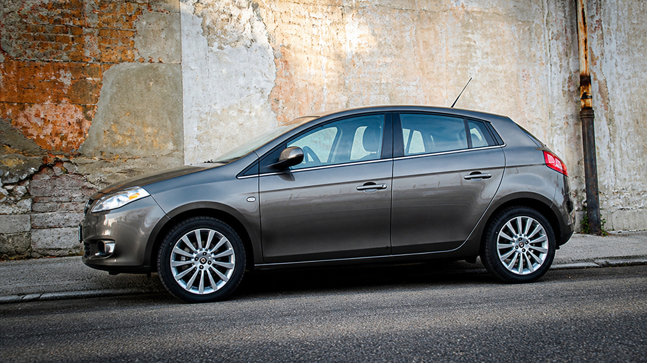 Grey five-door hatchback car parked beside a weathered brick and plaster wall on a street.