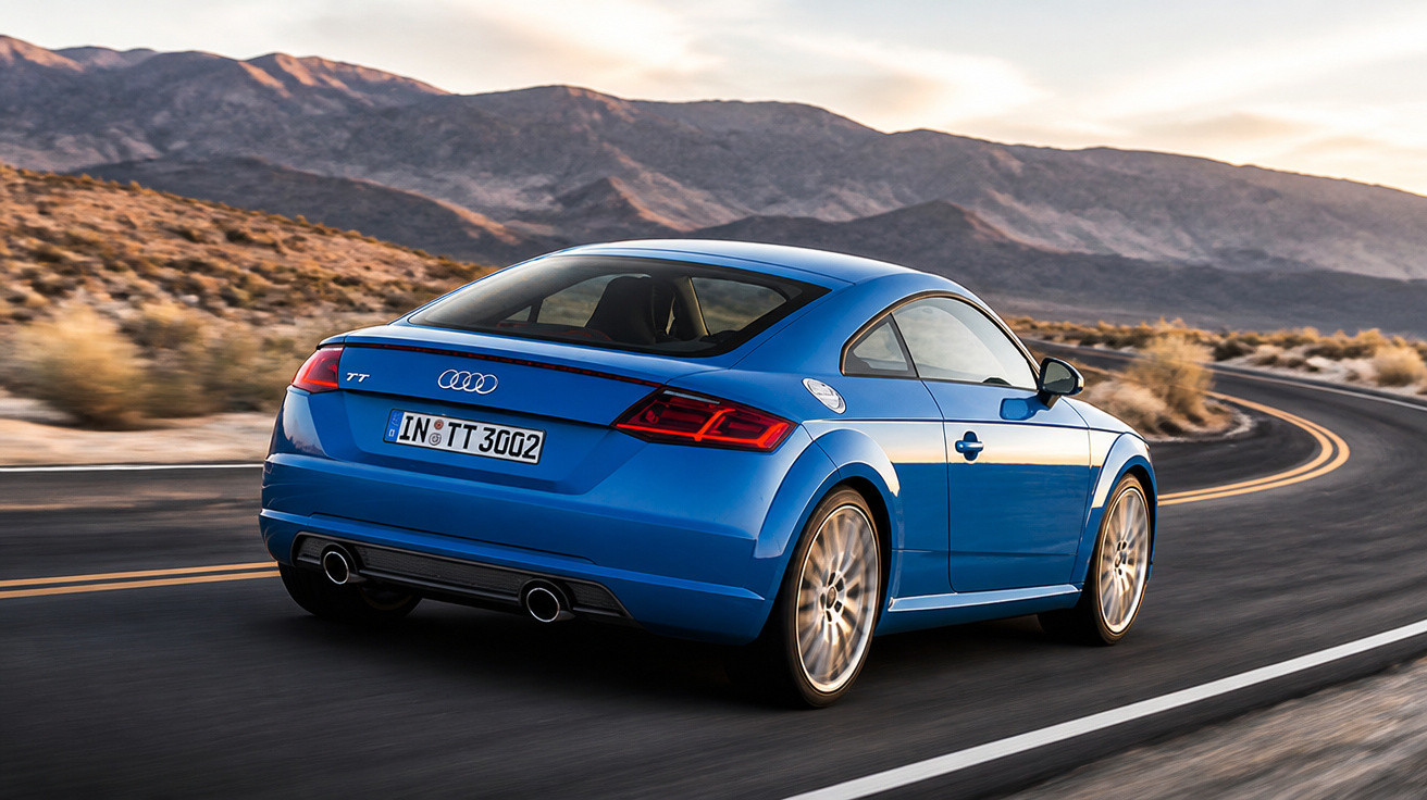 Blue Audi TT driving on a winding desert road with mountains in the background at sunset.