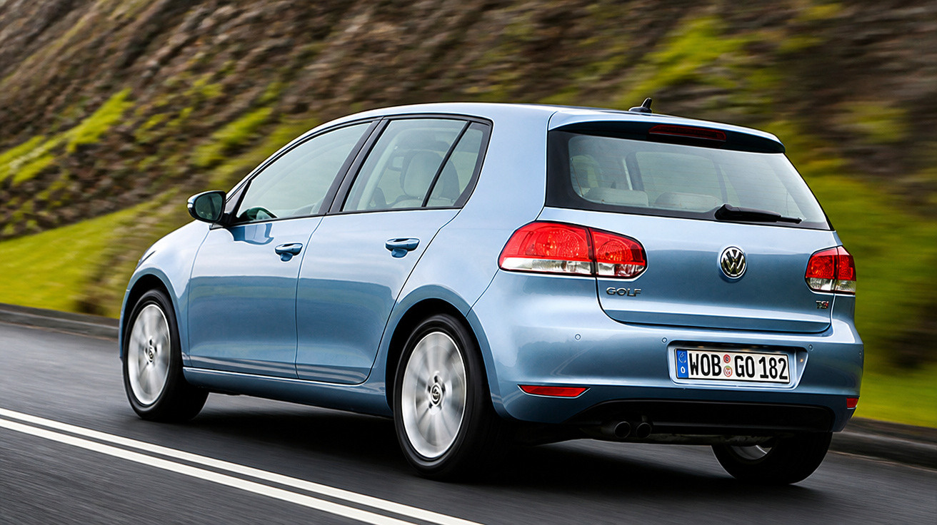 Blue Volkswagen Golf hatchback driving on a wet road with greenery and rocky hill in the background.