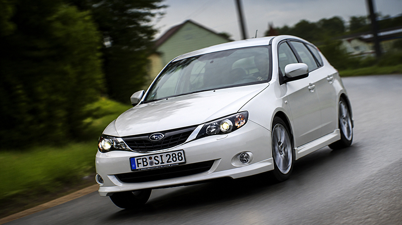 White Subaru hatchback driving on a wet road with blurred greenery and houses in the background.