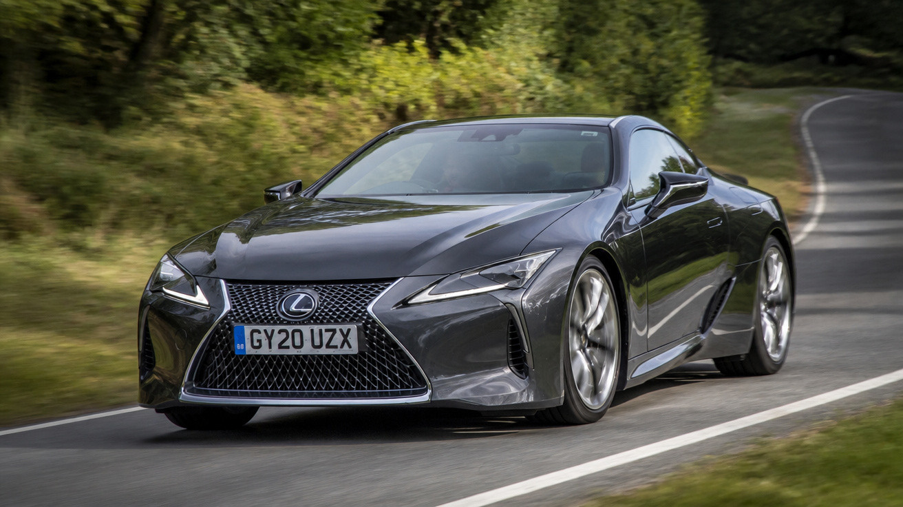Grey Lexus sports car driving on a winding road with trees in the background.