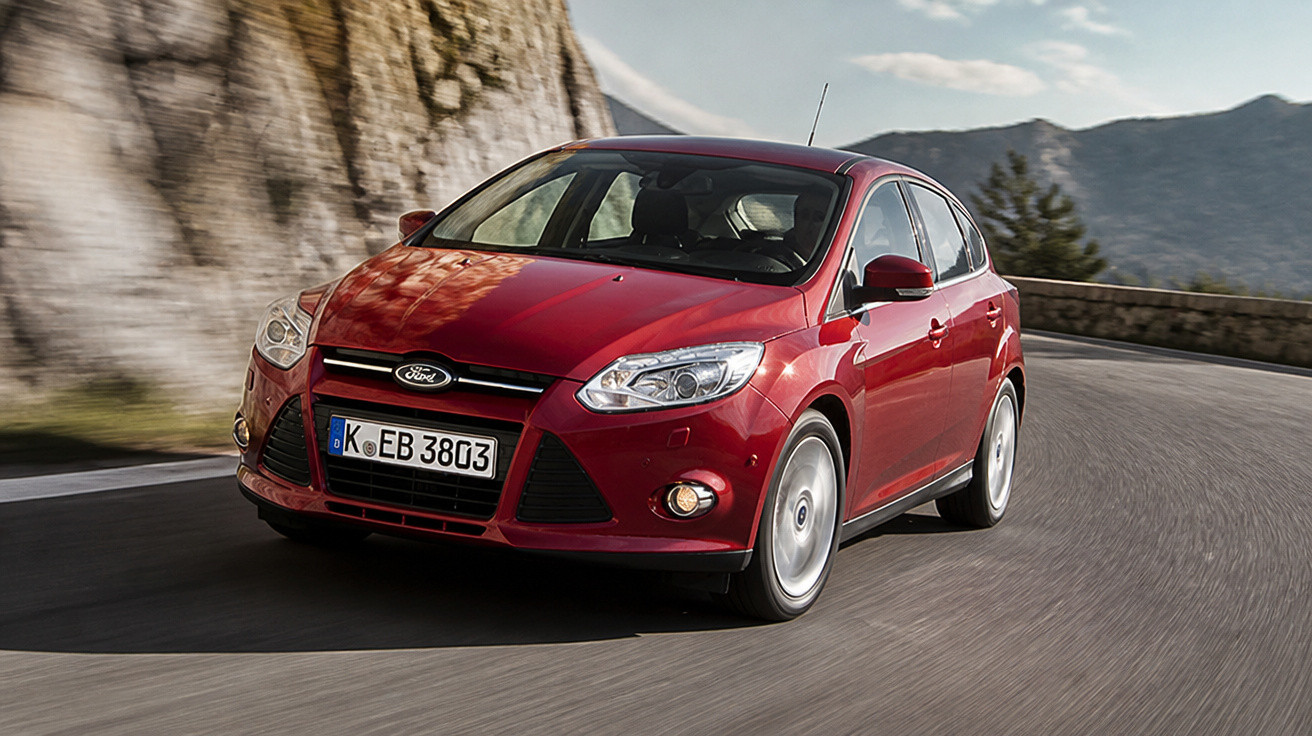 Red Ford hatchback car driving on a winding mountain road beside rocky cliffs.