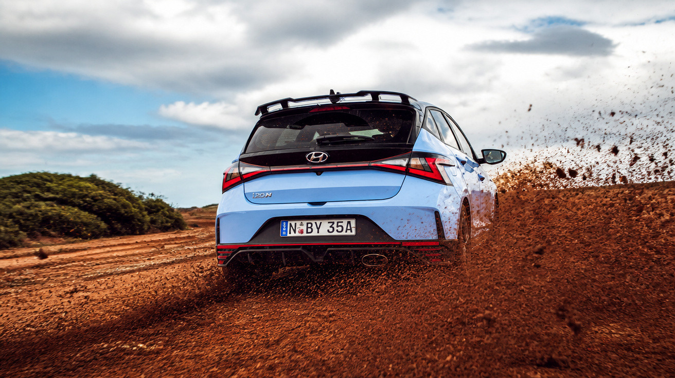 Blue Hyundai i20N car driving fast on dirt road, kicking up mud under cloudy sky