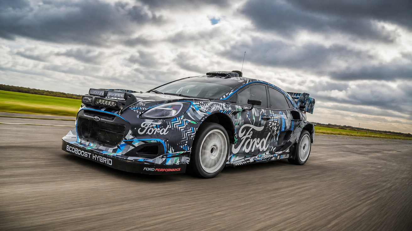 Blue and black Ford EcoBoost hybrid rally car speeding on a road under a cloudy sky.