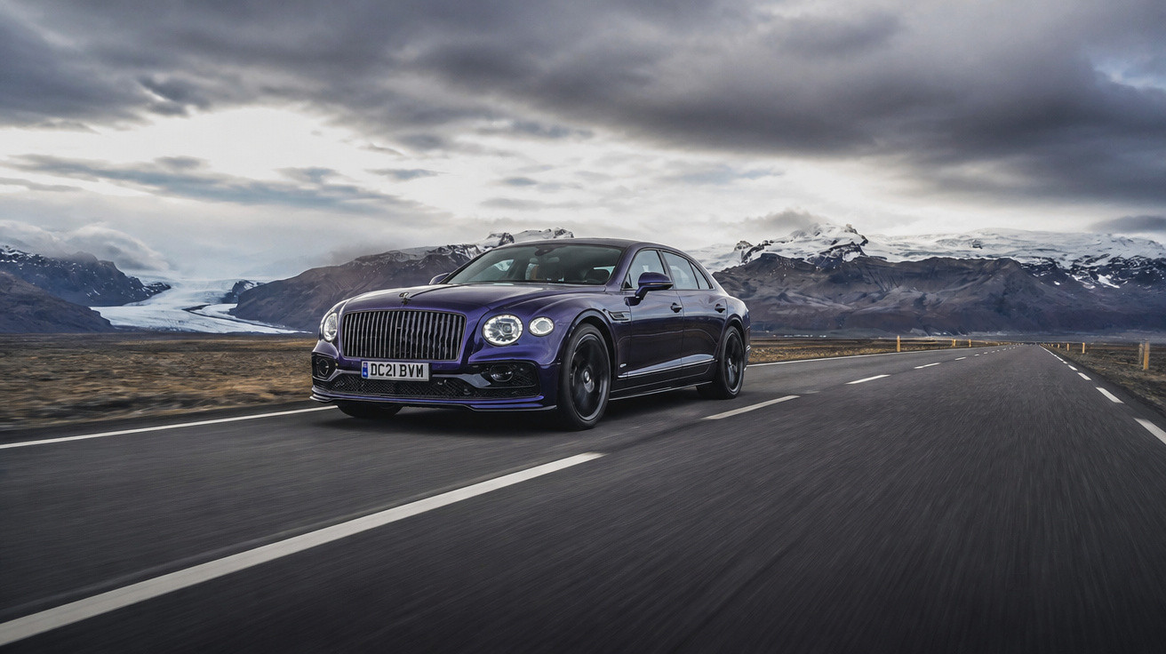 Black luxury sedan driving on empty road with snowy mountains and cloudy sky in the background.