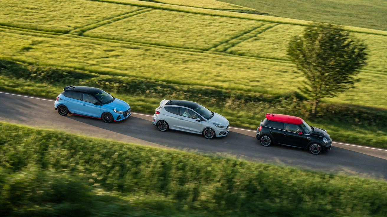 Three compact cars in blue, white, and black with a red roof driving on a rural road surrounded by green fields.