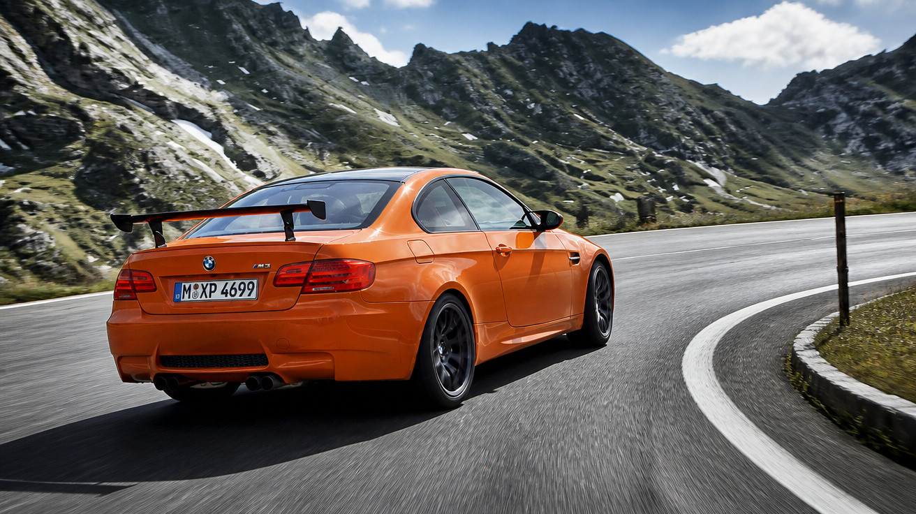 Orange BMW sports car driving on a winding mountain road with rocky peaks in the background.