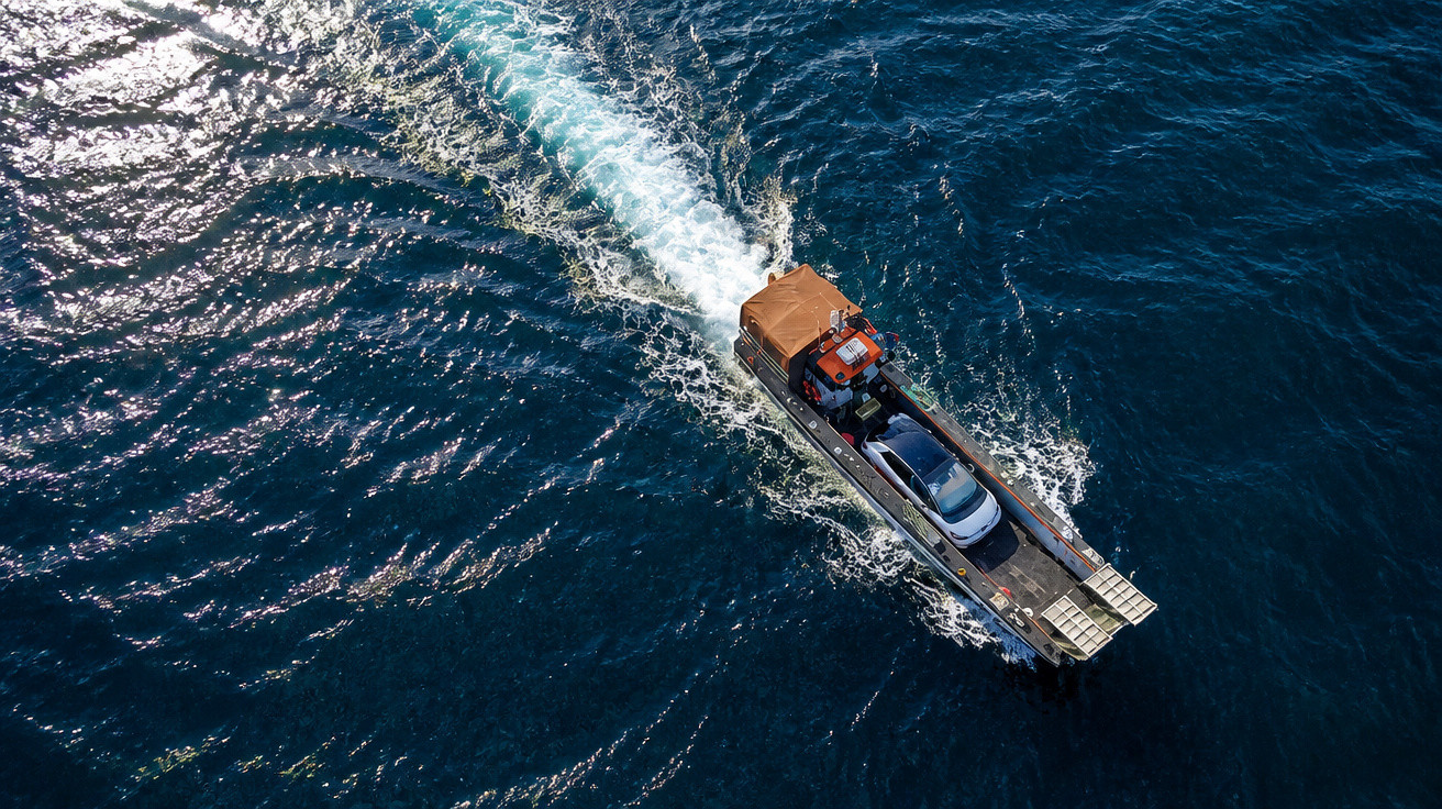 A small boat carrying a car moves through deep blue ocean water creating a white wake behind it.