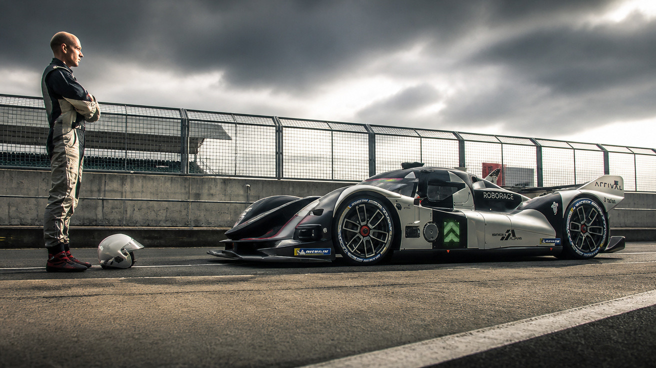 Racing driver in suit standing on pit lane, facing futuristic black and silver race car under cloudy sky.