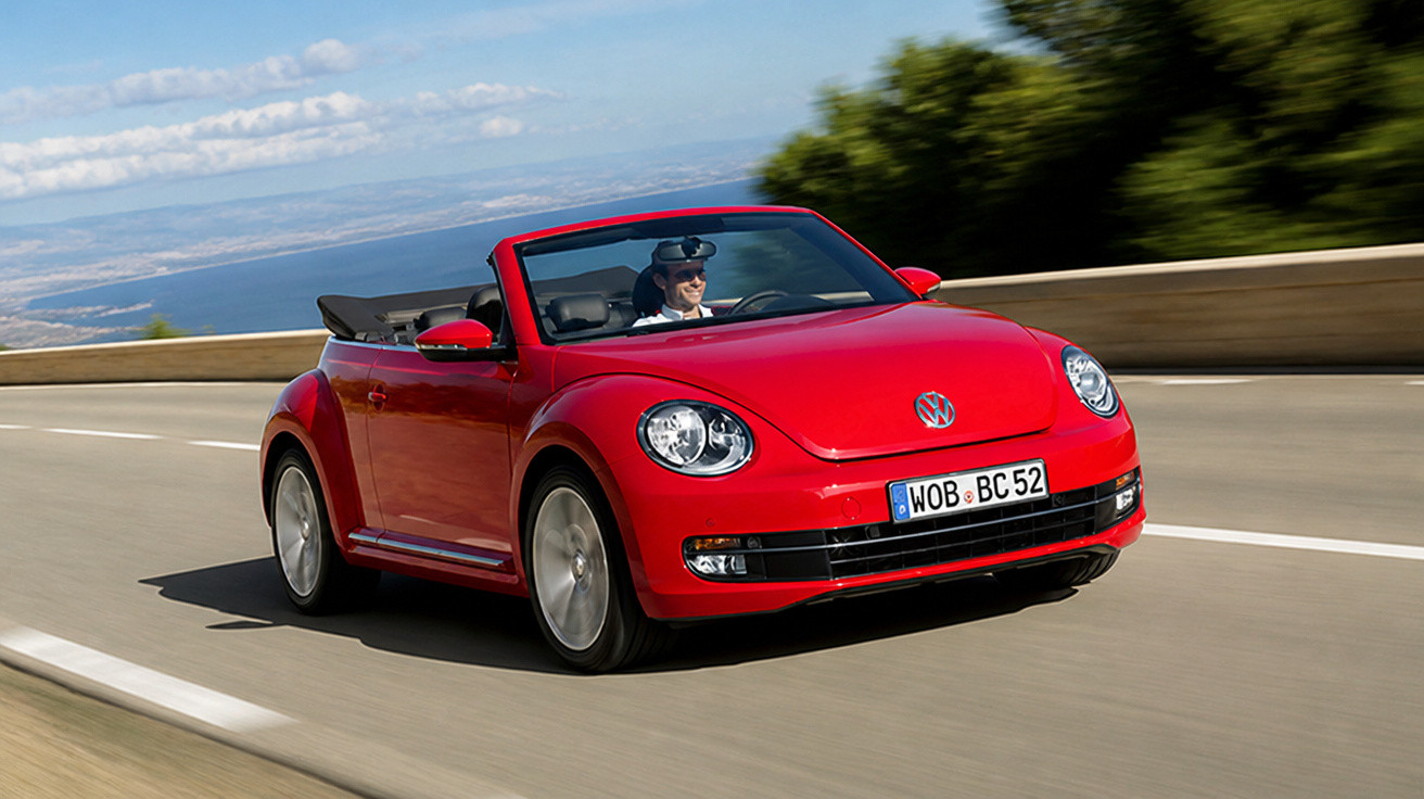 Red Volkswagen Beetle convertible driving on a coastal road with a driver wearing sunglasses.