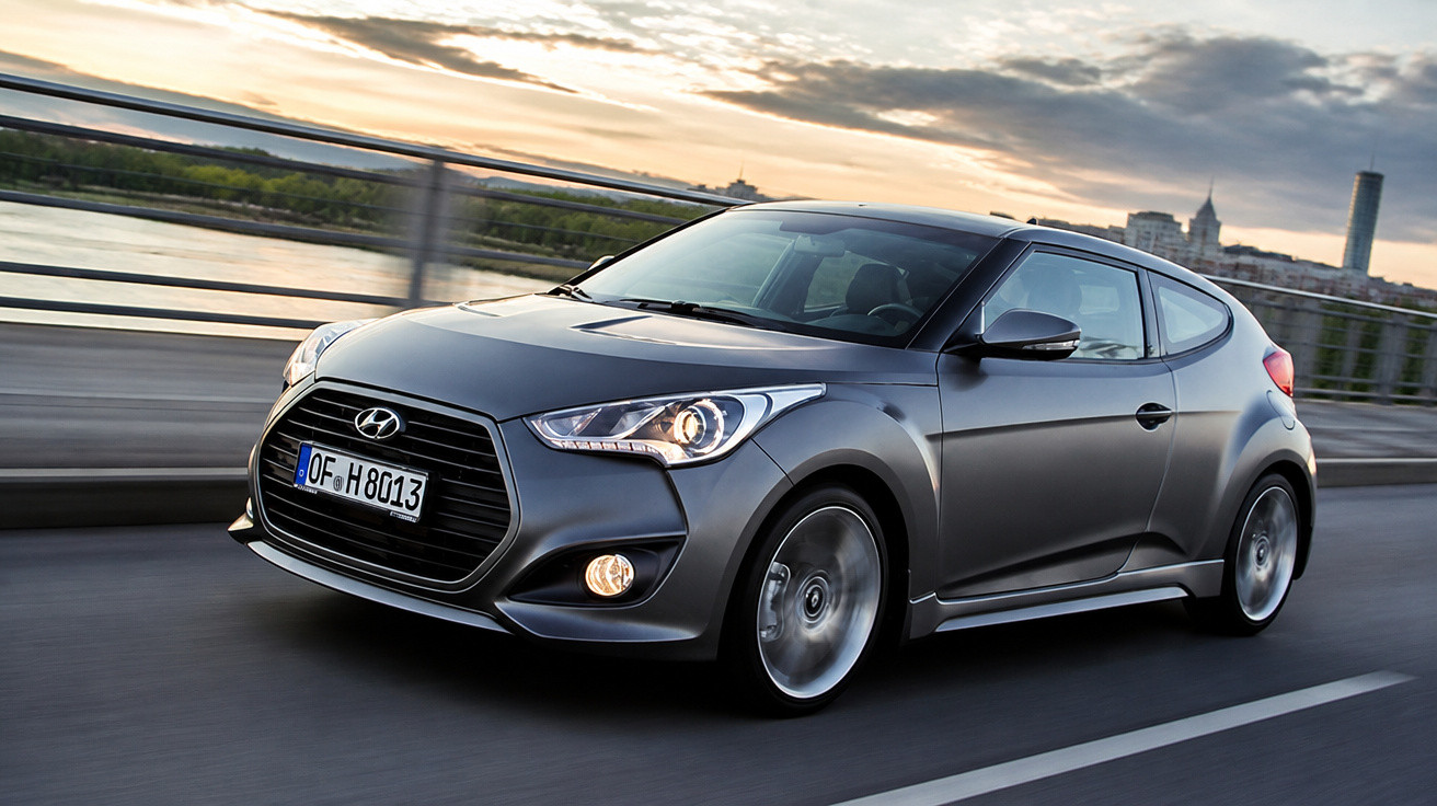 Grey Hyundai Veloster driving on a city bridge at sunset with blurred background and headlights on.
