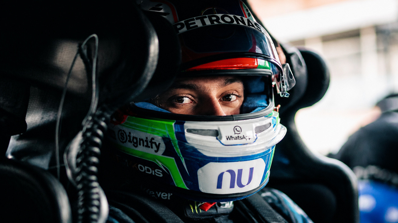 Close-up of a race car driver wearing a helmet with Petronas and WhatsApp logos, seated in the car cockpit.