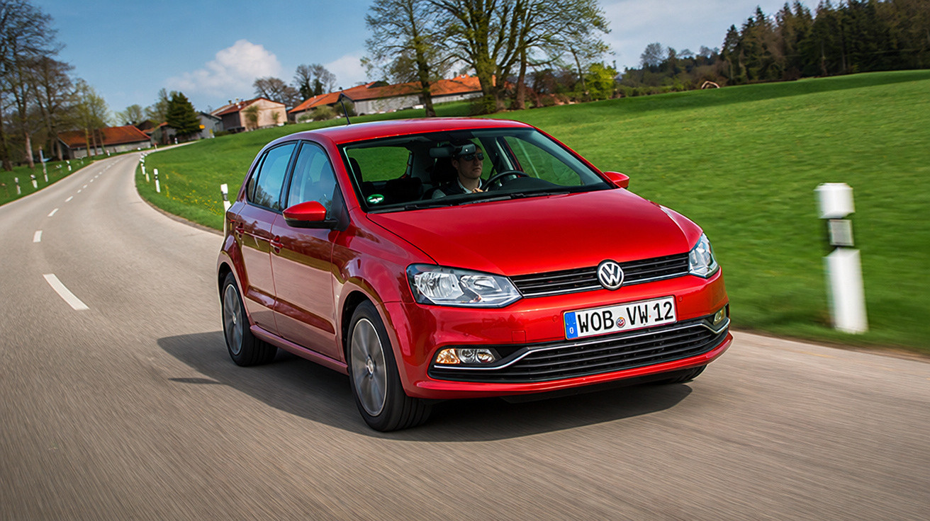 Red Volkswagen hatchback driving on a winding country road with green fields and houses in the background.