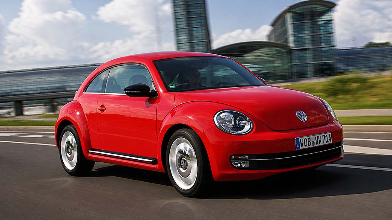 Red Volkswagen Beetle driving on a road with modern glass buildings in the background under a partly cloudy sky.