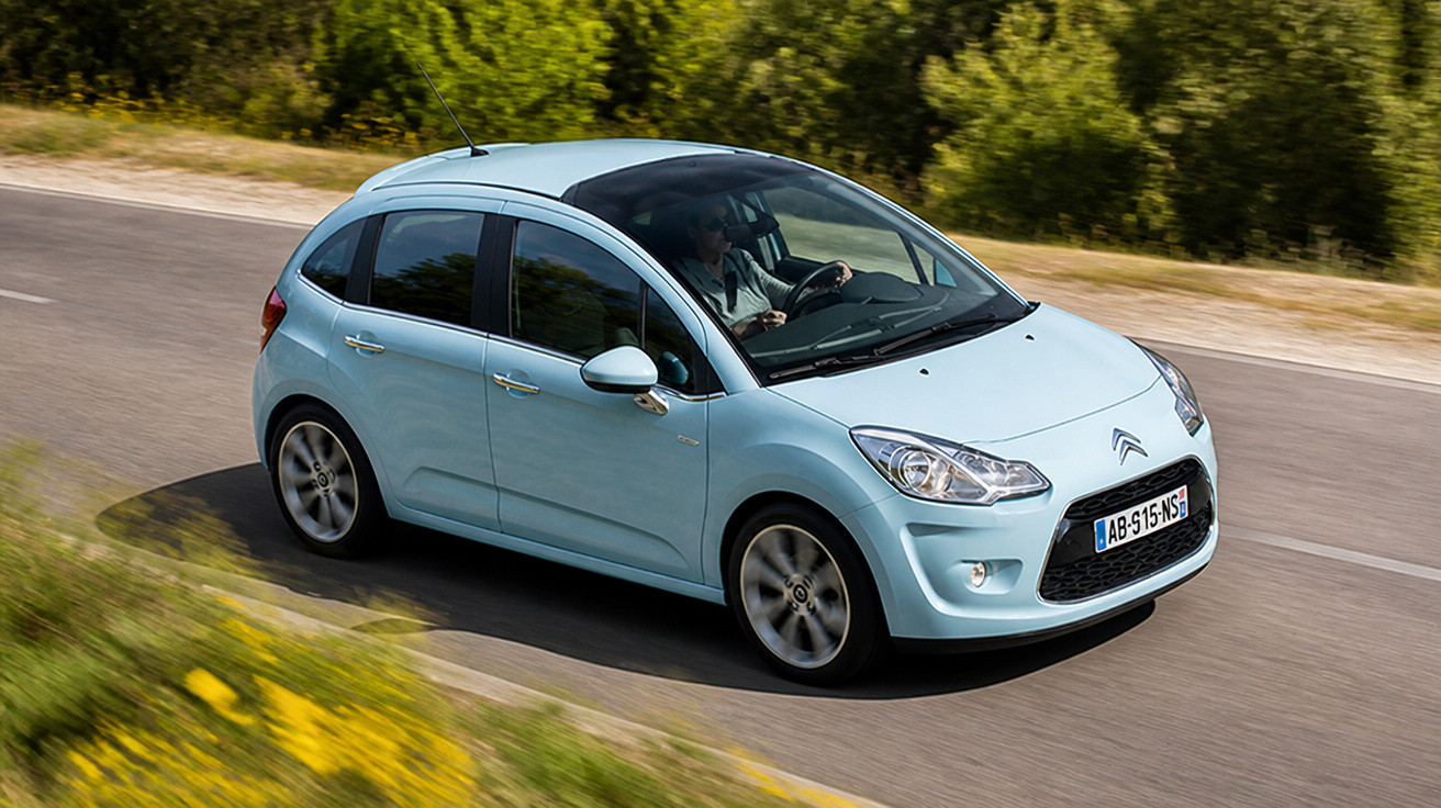 Light blue Citroën hatchback driving on a curved road surrounded by greenery and yellow flowers.