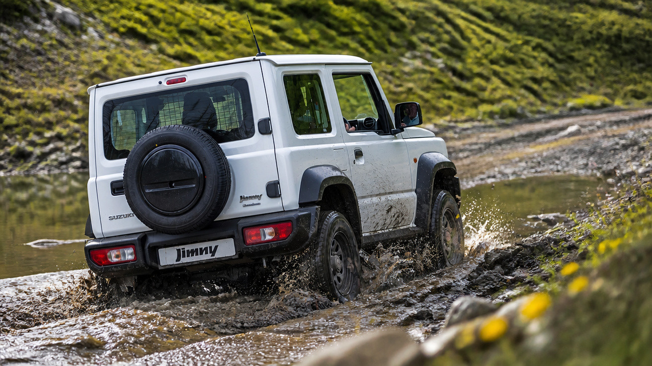 White Suzuki Jimny 4x4 off-road vehicle driving through muddy water on a rugged trail.