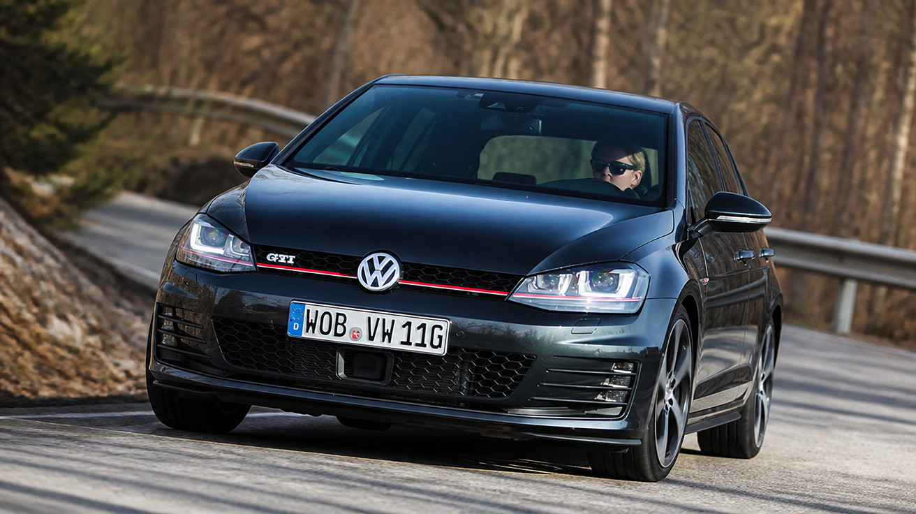 Black Volkswagen Golf GTI driving on countryside road with driver wearing sunglasses.